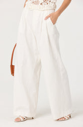 The image shows a person wearing high-waisted, wide-leg white pants with a relaxed fit. The pants have pleats at the waist and side pockets. The person is also wearing a white, sleeveless, crochet top with floral patterns. They have their right hand in their pocket and are wearing white strappy high-heeled sandals. A brown handbag with a thin strap is partially visible hanging on the person's left side.